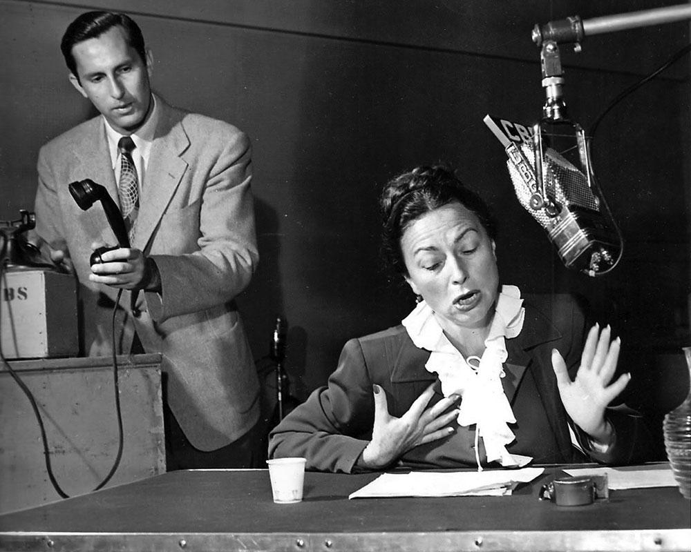 Black-and-white photograph of a 1950s radio drama studio: an actress performing at a table while a producer holds a telephone handset, under a hanging CBS microphone